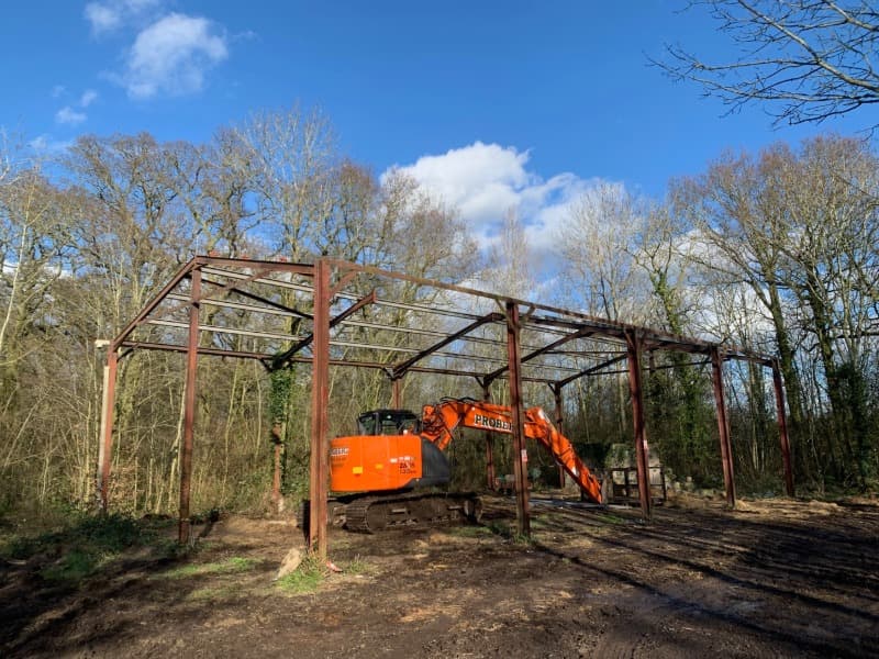 Steel framed farm barn with no roof, sides or ends. Just the frame stands over an orange excavator. The barn is in a woodland setting during winter. 