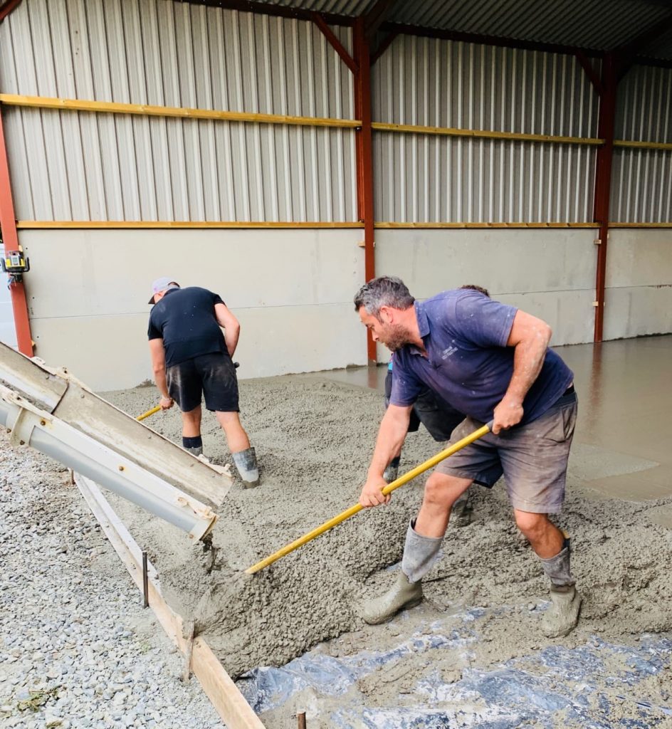 Two men in wellington boots stand in a barn as a lorry pours liquid concrete to make a smooth floor. The men rake the concrete into place. 