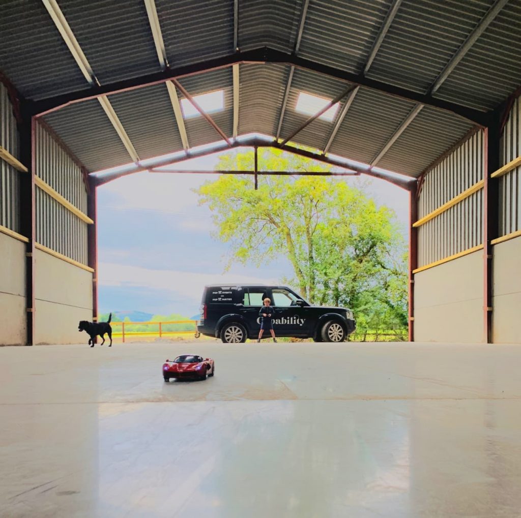 A boy is silhouetted against the open end of a storage barn. He is playing with a toy Ferrari car. A black dog runs after the car. 