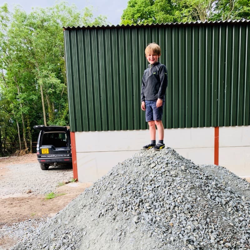A boy stands stands on top of a large pile of finishing stones outside a barn in the summer.