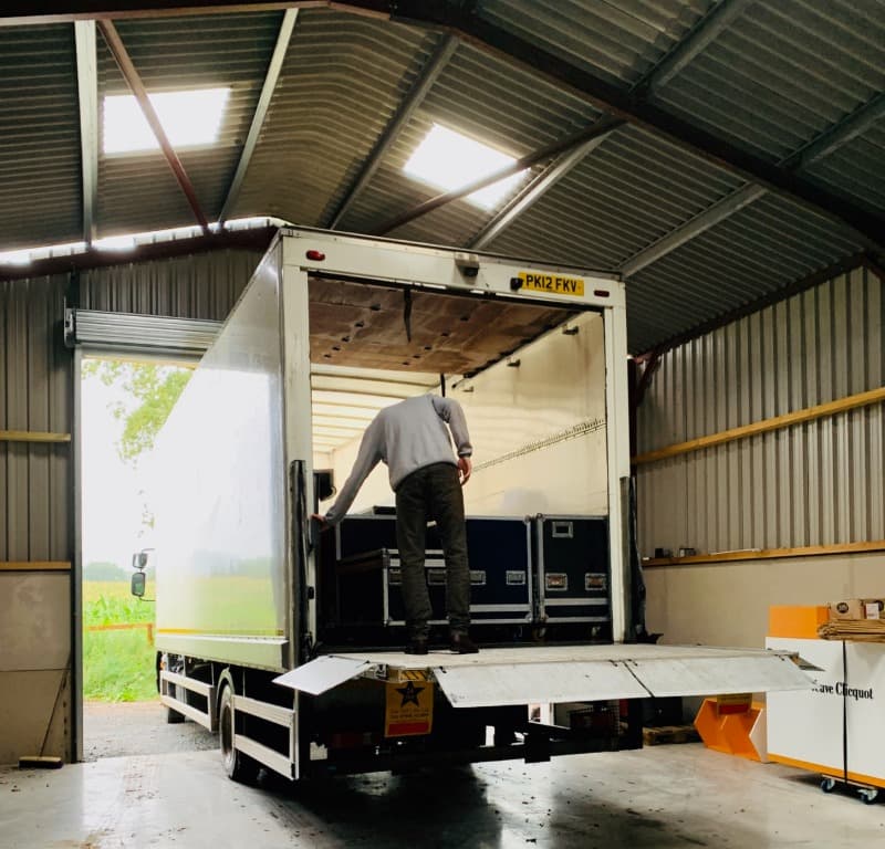 A large white lorry is backed into a storage barn roller shutter door. A man is unloading the lorry via it's tail lift. 