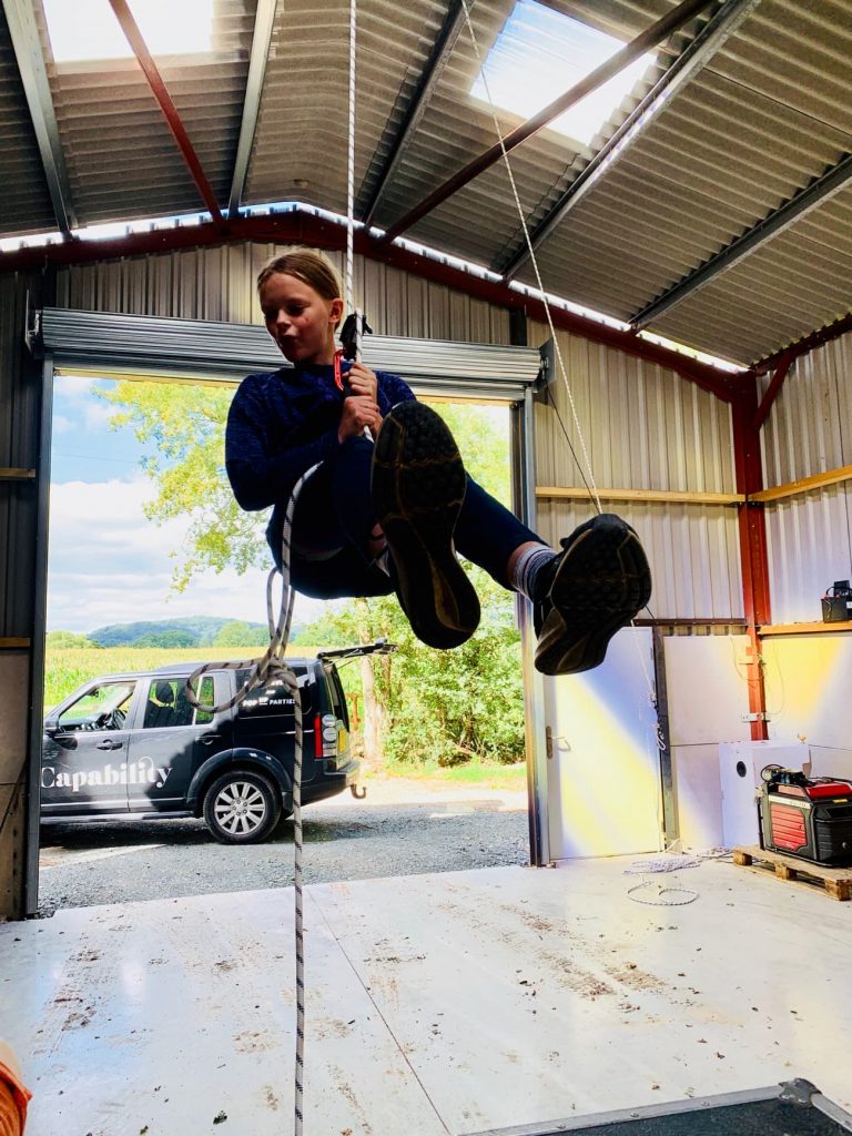 A girl climbs ropes into the roof of a storage barn in front of an open roller shutter door. A Land Rover Discovery is parked in the doorway. 