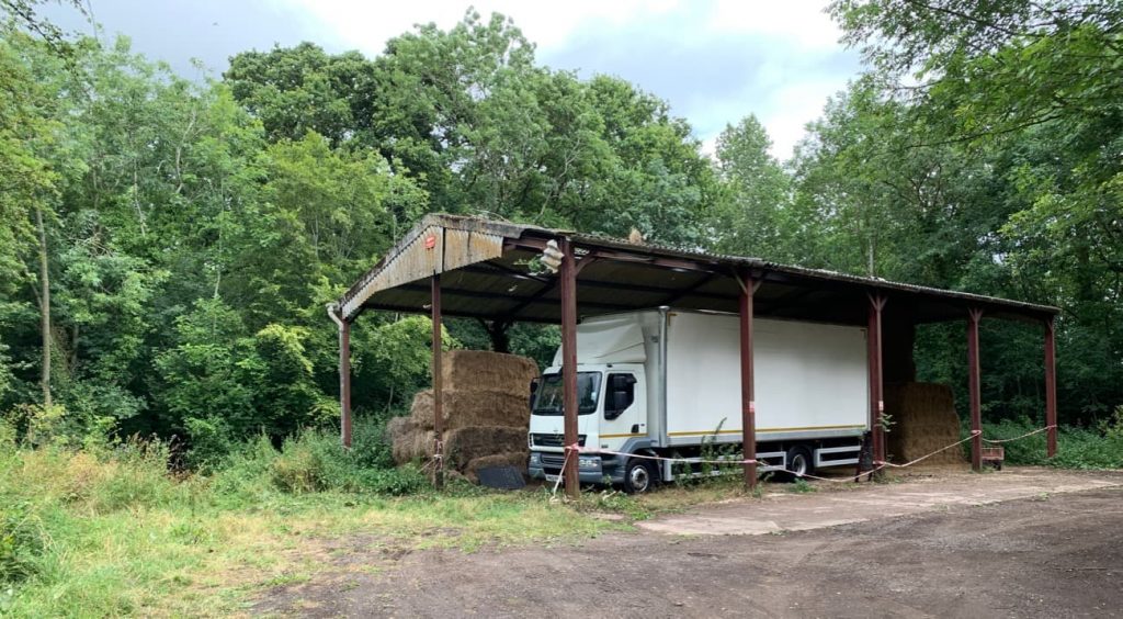 Farm barn with no sides in a wood during the spring. A white lorry sits inside the barn